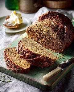 Slow Cooker Irish soda bread on green cutting board showing dense, rustic texture with herbs, butter and cream in background.